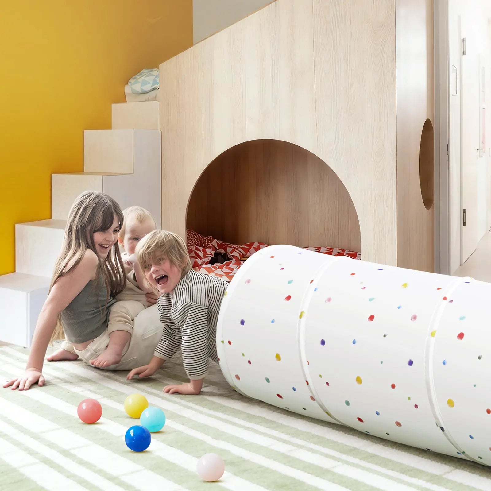 Three children play and laugh on a striped rug near a small indoor playhouse with stairs. Colorful balls and the Tiny Land® Adventure Play Tunnel by Tiny Land, featuring playful white spots, are scattered on the floor, creating a lively and fun atmosphere.|Colorful Dots