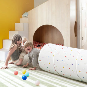 Three children play and laugh on a striped rug near a small indoor playhouse with stairs. Colorful balls and the Tiny Land® Adventure Play Tunnel by Tiny Land, featuring playful white spots, are scattered on the floor, creating a lively and fun atmosphere.|Colorful Dots