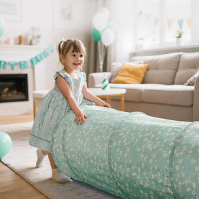 A young girl in a blue floral dress smiles and plays with the Tiny Land® Adventure Play Tunnel - Green by Tiny Land in a bright living room decorated with balloons, a couch, and a birthday banner.|green