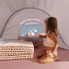 A young girl sits inside a Tiny Land® One-Stop Beach Toy & Tent Set – Blush Pink, gazing at the ocean. Woven mats are stacked nearby and sand covers the tent floor, highlighting this UV-protective set by Tiny Land.