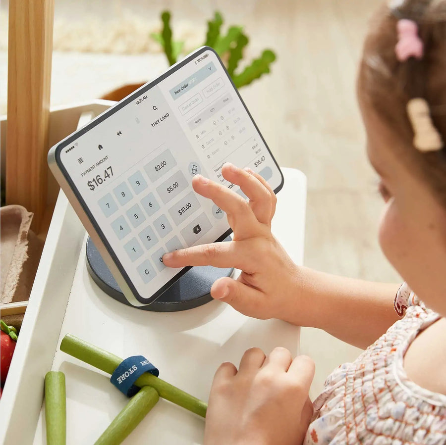 A child plays pretend with the Tiny Land® Kid's Grocery Store Playset, using a touchscreen tablet as a cash register with a calculator app, alongside vegetables and a toy receipt for realistic shopping fun.