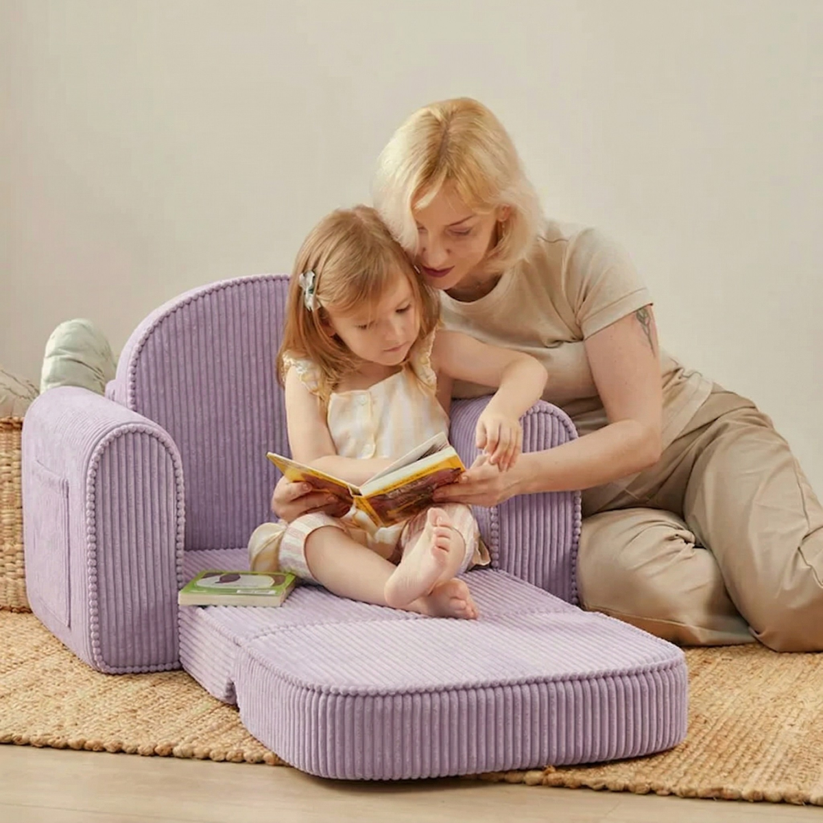 A woman and a young girl sit together on the floor, reading. The girl relaxes on the Tiny Land® GentleSnug Kid Chair in purple, while the woman sits beside her and helps guide her through the book.