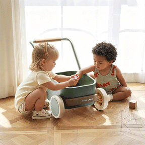 Two young children sit on the floor by a Tiny Land® JoyRide Baby Walker with Blocks - Sage from Test. They play together in natural light, blocks scattered around them, with a bright window in the background.