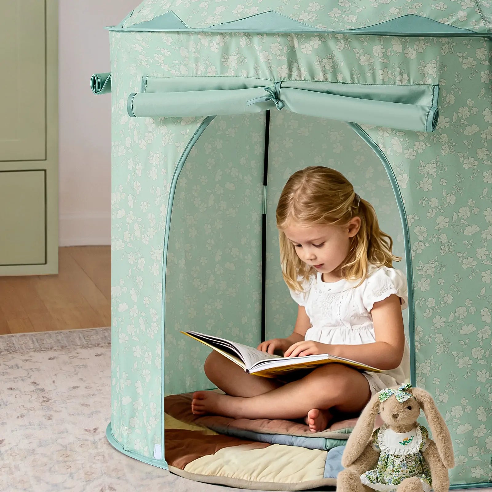 A young girl in a white dress reads inside the Tiny Land® Tiny Sprout Castle Tent by Tiny Land, sitting cross-legged on a quilted mat with a stuffed bunny nearby—a cozy and peaceful indoor scene.
