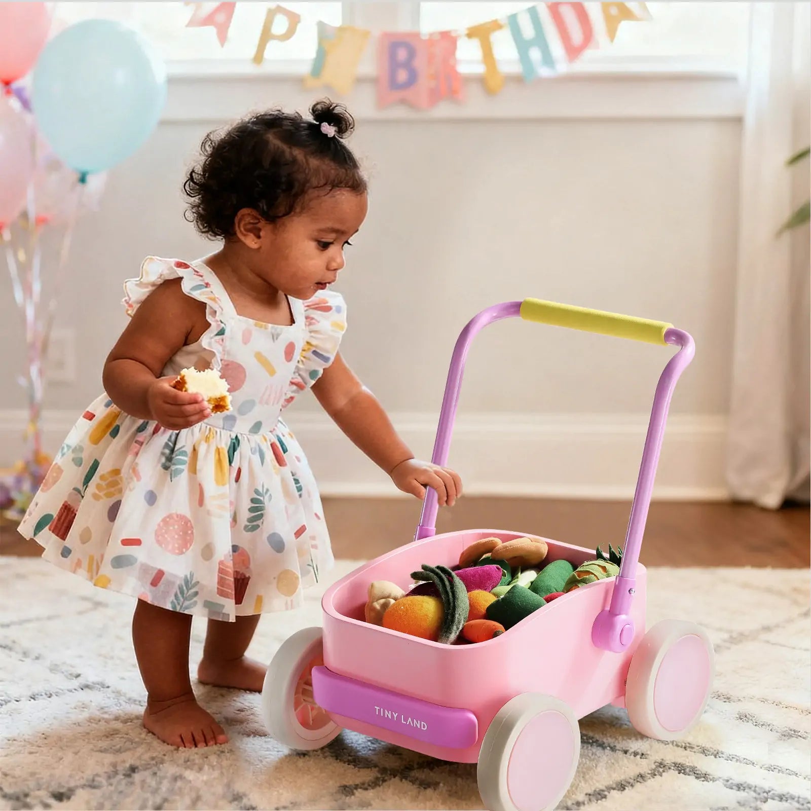 A toddler in a colorful dress holds a toy cookie and plays with the Tiny Land® 3-in-1 Wooden Baby Walker Wagon - Pink, filled with play food. Birthday balloons and decorations from Tiny Land are visible in the background.