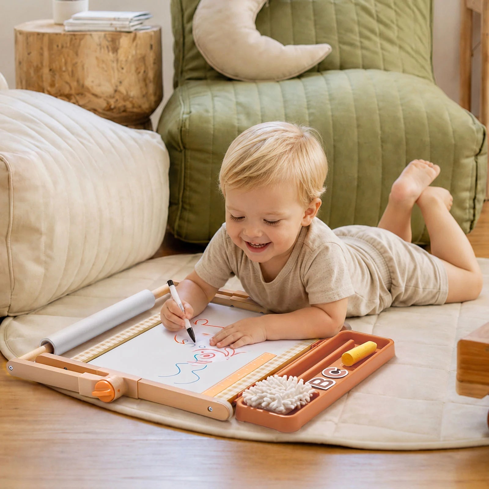 A smiling toddler lies on a mat, drawing with colorful markers on the Tiny Land® Double-Sided Magnetic Tabletop Easel. Cozy cushions and wooden decor from Tiny Land set the perfect scene for a playful kids’ art station.|Oatmeal