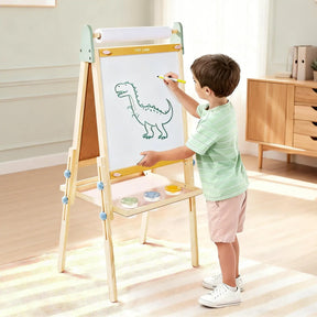 A young boy draws a dinosaur on the white side of a Tiny Land® Morandi 3-in-1 Art Easel & Paper Roll Set, which features an adjustable height, a paper roll at the top, and paint cups on a tray.