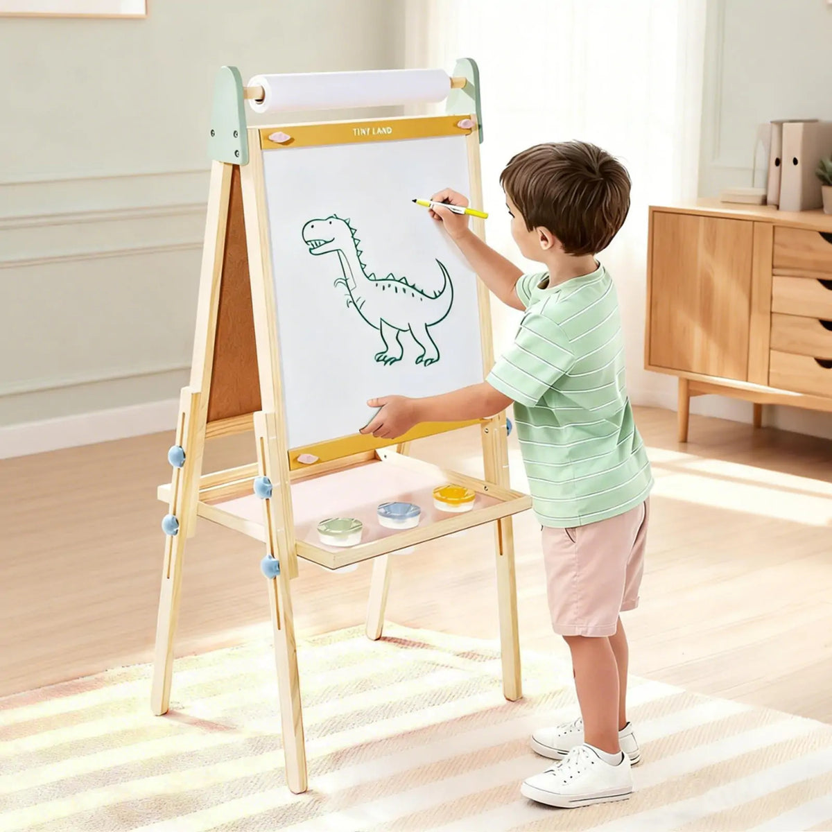 A young boy uses the Tiny Land® 3-in-1 Art Easel for Kids - Morandi, drawing a dinosaur on white paper with a yellow pencil in a bright, tidy room. Three paint pots rest on the shelf below the easel from Tiny Land.