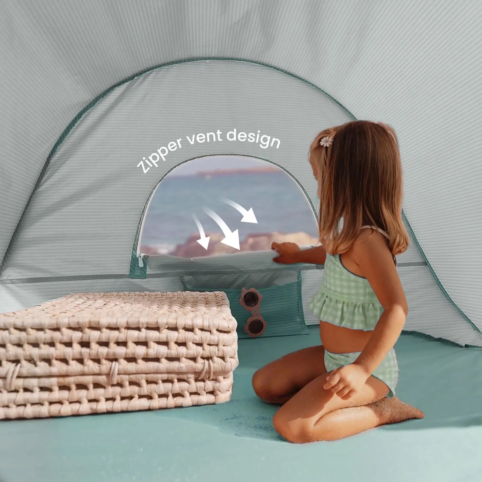A young girl in a green swimsuit sits inside the Tiny Land® One-Stop Beach Toy & Tent Set – Sea Blue, peering through its zipper vent window as she enjoys the beach view next to two woven mats.