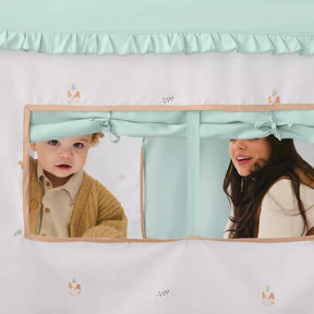 A baby in a brown sweater and a woman with long dark hair peek through the Tiny Land® Sweet Woodland Kids Play Tent by Tiny Land INC, featuring white and pastel colors, ruffled trim, decorative patterns, and a cozy play mat inside.
