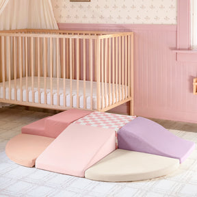 A wooden crib stands in a pastel pink nursery next to the Tiny Land® PlayNest Climbing Blocks—foldable foam play set pieces by Tiny Land in pink, purple, beige, and pink-checkered patterns, arranged flower-like on a light rug.
