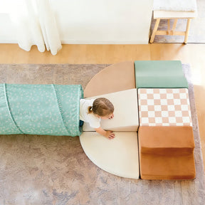 A young child crawls out of the Tiny Land® Adventure Play Tunnel - Green onto a soft play mat with colorful foam blocks arranged in a semi-circle on a patterned rug.|green
