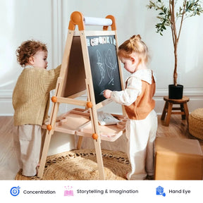 Two young children draw with chalk on a standing Tiny Land® Caramel Magnitales Easel for Kids in a cozy, sunlit room. This double-sided magnetic easel by Tiny Land encourages creative play from every angle, while educational icons and text below highlight key skills: concentration, storytelling & imagination, and hand-eye coordination.