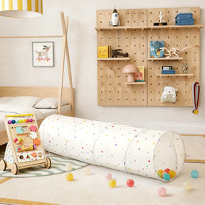 A cozy, modern children's room includes a wooden bed, pegboard shelves with toys and books, the Tiny Land® Adventure Play Tunnel - Colorful Polka Dots, a wooden activity walker, and scattered plastic balls on a light rug.