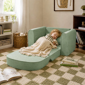 A small child naps on the Tiny Land® GentleSnug Kids Sofa Chair in a cozy room, covered with a beige blanket and holding a stuffed animal. Books and toys are scattered around as sunlight streams through the window.