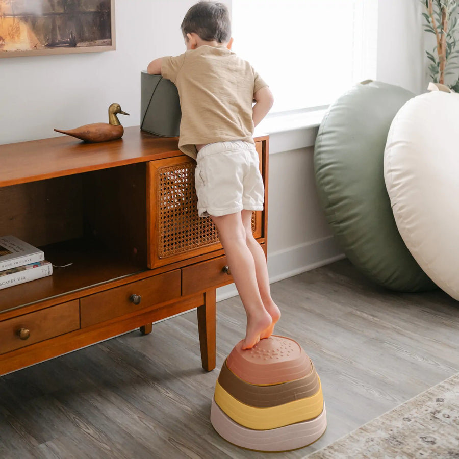 A young child uses Tiny Land® Stepping Stones - Sunset Hues by Tiny Land to build balance and coordination while reaching for a box atop a wooden cabinet in a bright, modern living room.|Sunset Hues