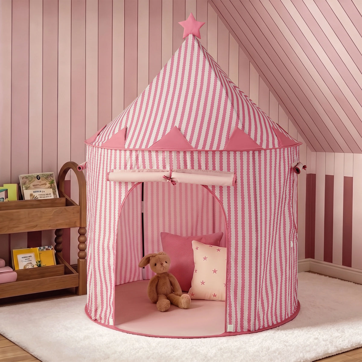 A Tiny Land® Kids Tent – Indoor Tent for Toddlers by Tiny Land, featuring pink and white stripes with a star on top, sits on a white rug. Inside the tent are a brown stuffed bunny and a pink pillow. Nearby, a wooden shelf displays children’s books. The room’s pink striped and paneled walls create the perfect setting for this charming indoor play tent or playhouse for kids.|PInk