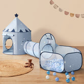 The Tiny Land® Adventure 3-in-1 Play Tunnel & Ocean Ball Set - Blue features a blue castle-shaped tent with stars, a connected tunnel, and a ball pit with white and blue balls. A teddy bear sits on a mat nearby, beneath a garland on the beige wall.
