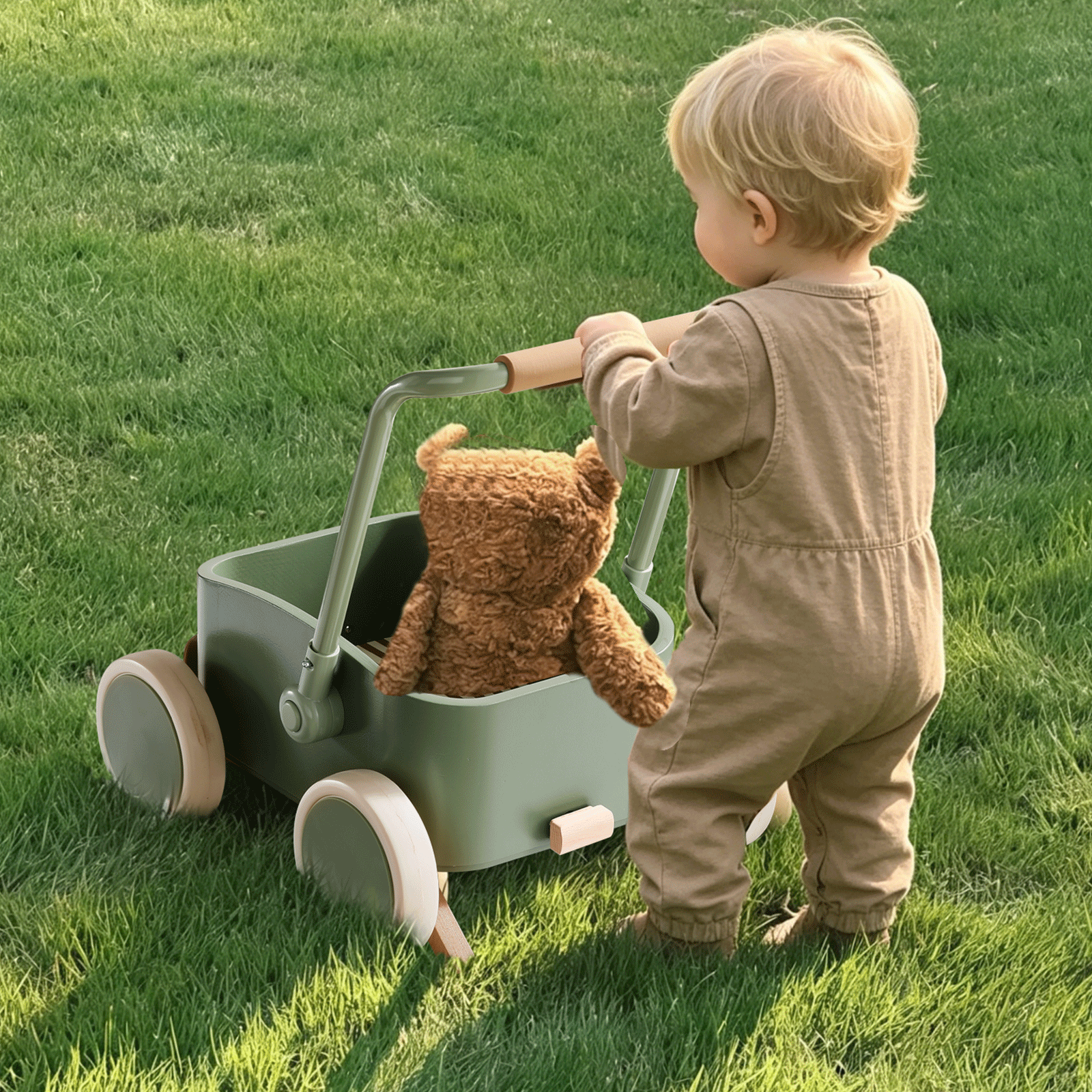 A toddler in a beige jumpsuit pushes the Test Tiny Land® JoyRide Baby Walker with Blocks - Sage, featuring adjustable speed control and a brown teddy bear inside, across a grassy lawn.