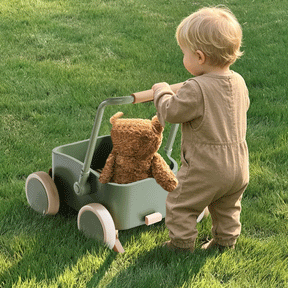 A toddler in a beige jumpsuit pushes the Test Tiny Land® JoyRide Baby Walker with Blocks - Sage, featuring adjustable speed control and a brown teddy bear inside, across a grassy lawn.