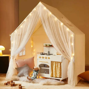 The Tiny Land® Chef’s Corner Playset combines a cozy cream playhouse with sheer curtains and string lights, a matching kitchen set, plush pillows, a stuffed mouse, wooden blocks, and a fluffy rug for endless pretend play.