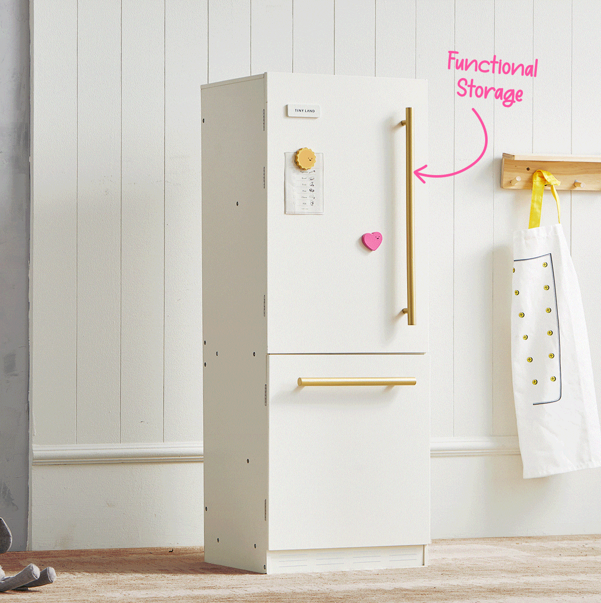 The Tiny Land® PopJoy Kitchen & Fridge Set features a white, modern toy fridge with gold handles. Perfect for pretend play, it stands against a paneled wall with an apron nearby and a pink heart magnet on the door labeled “Functional Storage.”.
