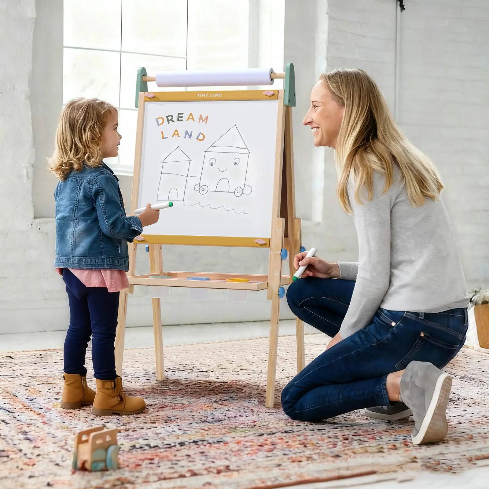 A young child and an adult smile together while drawing houses and “Dream Land” on the whiteboard of the Tiny Land® 3-in-1 Art Easel for Kids - Morandi by Tiny Land in a bright, cozy room.