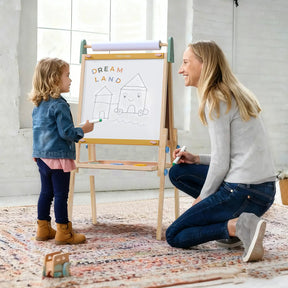 A young child and an adult smile together while drawing houses and “Dream Land” on the whiteboard of the Tiny Land® 3-in-1 Art Easel for Kids - Morandi by Tiny Land in a bright, cozy room.