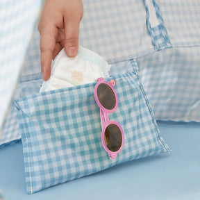 A hand puts a diaper into a blue gingham pouch with pink sunglasses, ideal for beach days with the Tiny Land® One-Stop Beach Toy & Tent Set – Light Blue Gingham. The matching pattern appears in the background.