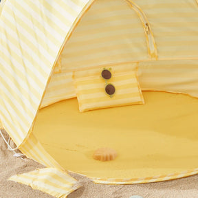 The Tiny Land® One-Stop Beach Toy & Tent Set in pastel yellow stripes sits on the sand, with a matching pillow holding round sunglasses and a seashell beneath its UV-protective shade—perfect for kids' beach fun.