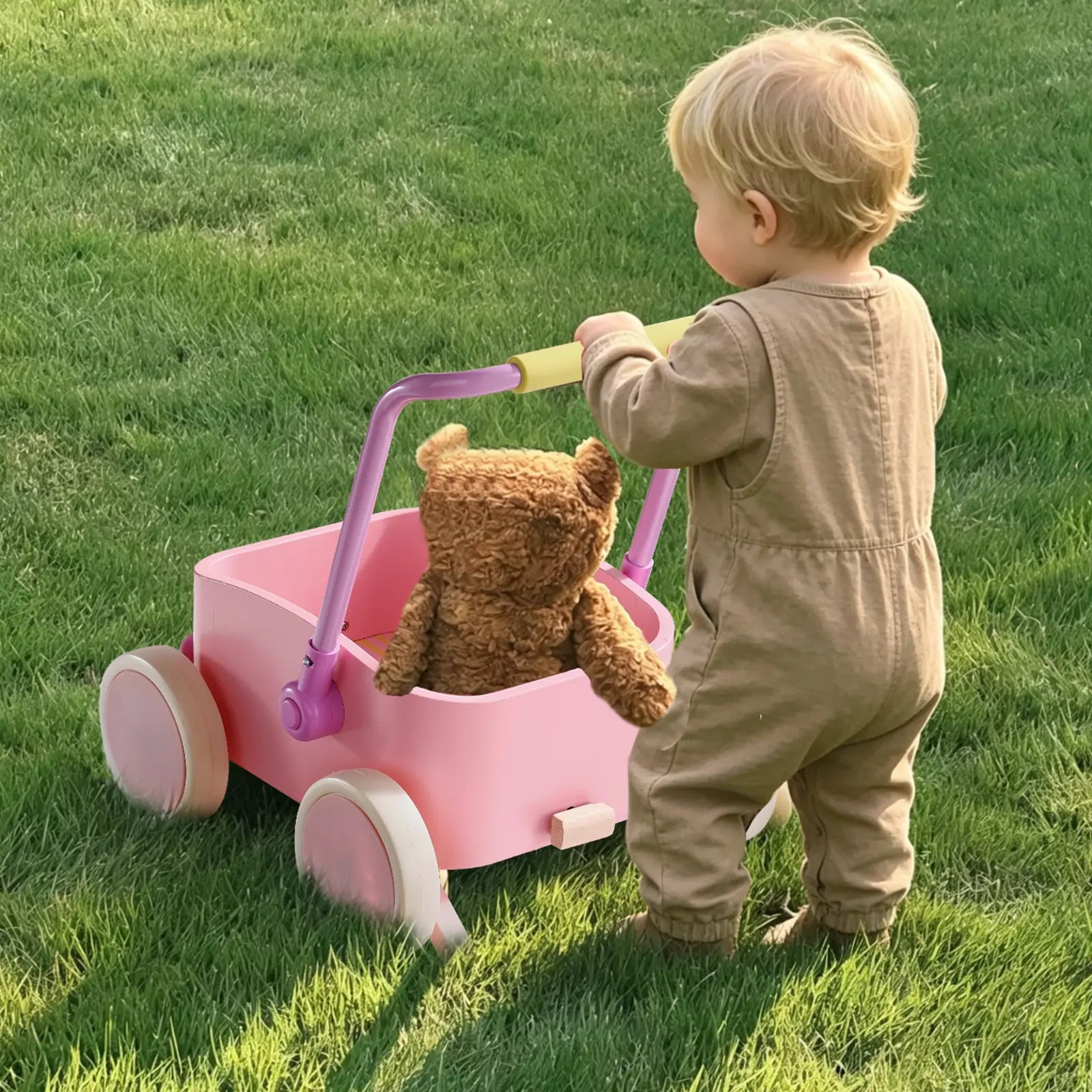 A toddler in a tan jumpsuit pushes the Tiny Land® 3-in-1 Wooden Baby Walker Wagon - Pink by Tiny Land, carrying a brown teddy bear, across a grassy lawn.