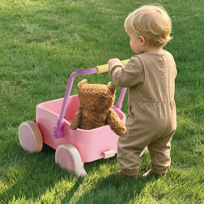 A toddler in a tan jumpsuit pushes the Tiny Land® 3-in-1 Wooden Baby Walker Wagon - Pink by Tiny Land, carrying a brown teddy bear, across a grassy lawn.
