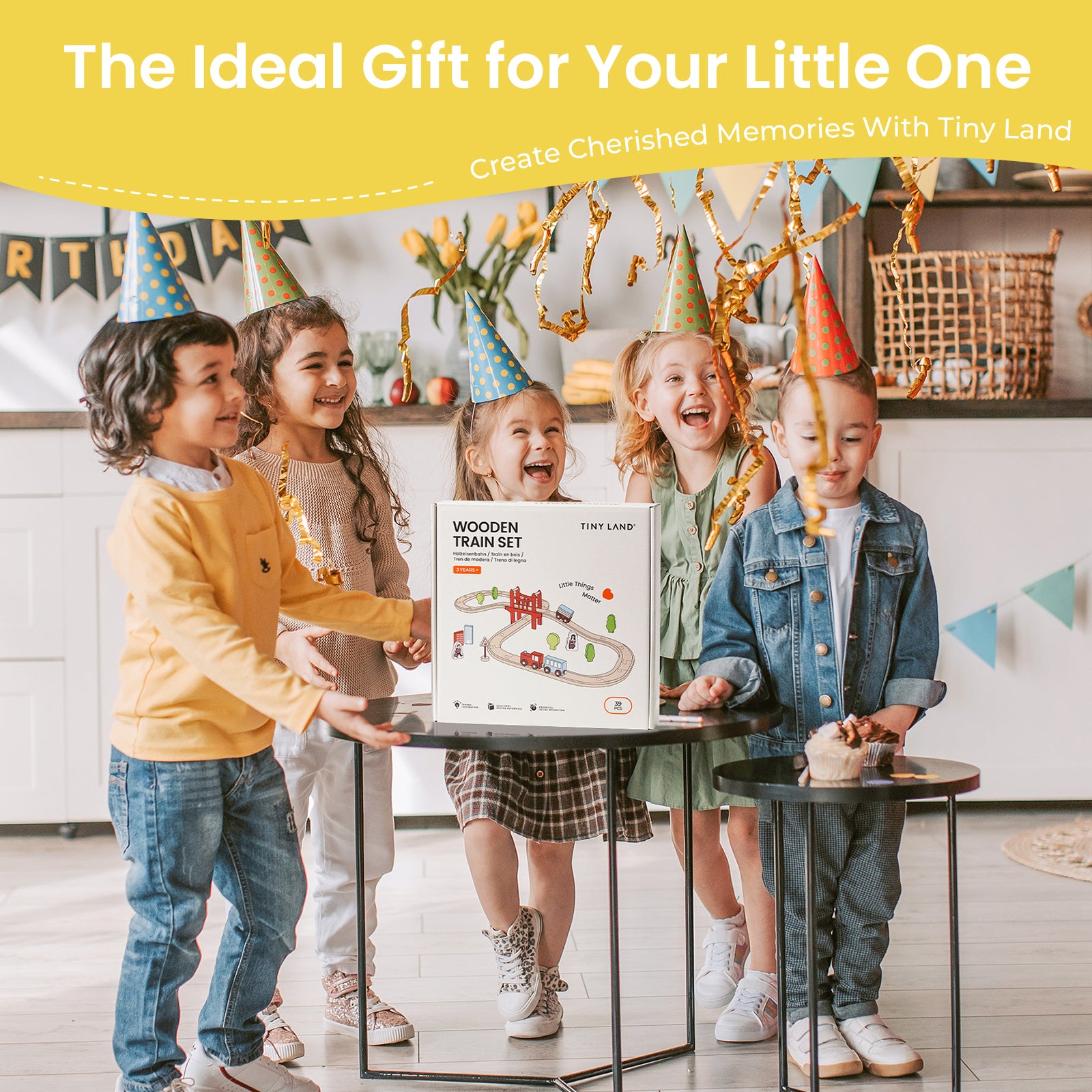 Four children in party hats smile around a gift box labeled "Tiny Land® Wooden Train Set for Children 39 Pcs" by Tiny Land, placed on a table. The festive background features colorful decorations and golden streamers.