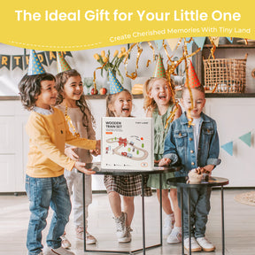 Four children in party hats smile around a gift box labeled "Tiny Land® Wooden Train Set for Children 39 Pcs" by Tiny Land, placed on a table. The festive background features colorful decorations and golden streamers.
