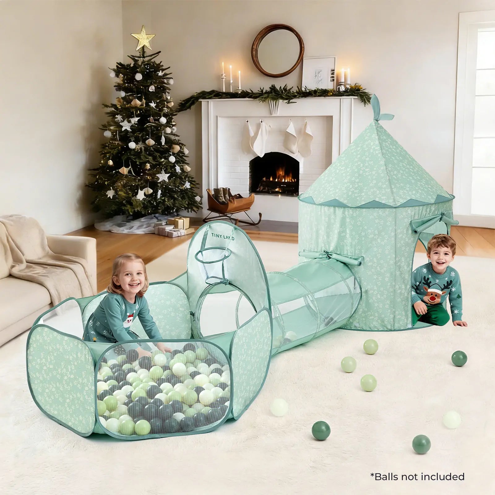 Two young children play in a Tiny Land® Compact Adventure 3-in-1 Play Tunnel With no Balls - Green, which features a cozy tent and tunnel. The living room is decorated for Christmas with a tree and fireplace, and some plastic balls are scattered on the carpet.