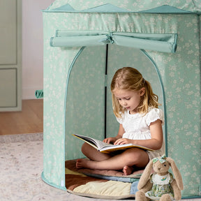A young girl sits cross-legged inside a pale green Tiny Land® Compact Adventure 3-in-1 Play Tunnel, reading. A stuffed bunny in a dress is on the rug beside her, while the play tunnel peeks out from one side of the tent.