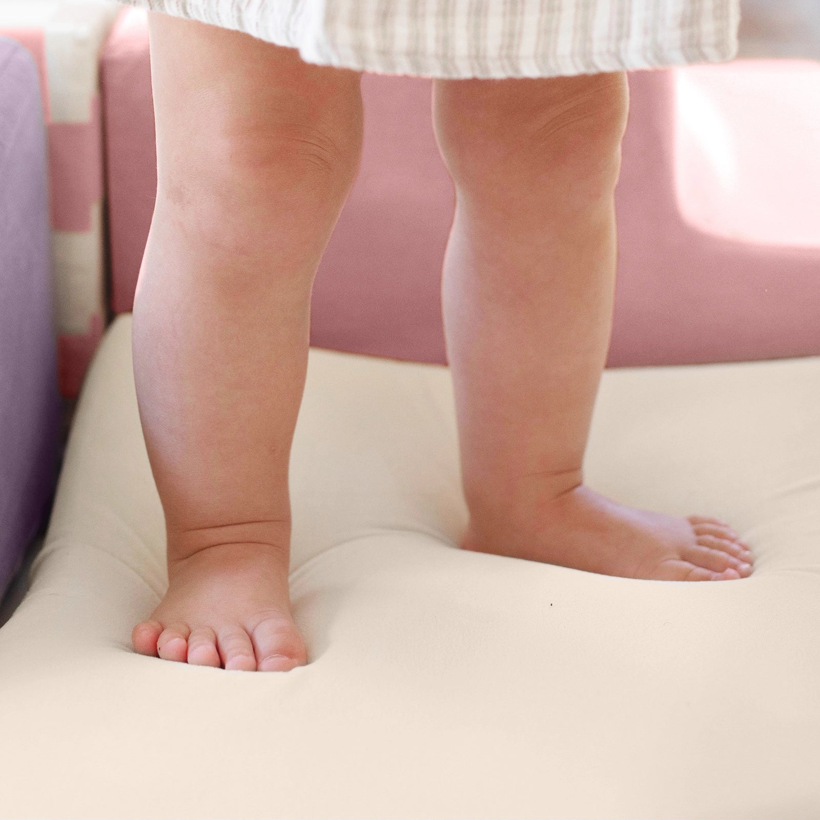 A young child stands barefoot on a soft, light-colored cushion, with only their legs and feet visible. They're wearing a striped dress, surrounded by Tiny Land® PlayNest Climbing Blocks for added fun. Brand: Tiny Land.