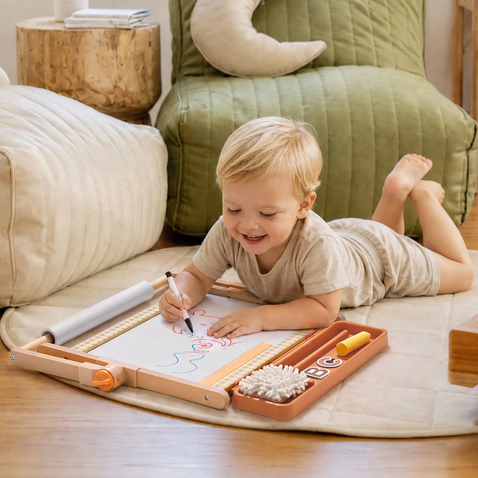 A young child lies on a soft mat, smiling as they draw with markers on paper at their Tiny Land® Double-Sided Magnetic Tabletop Easel. Art supplies are nearby, and cozy cushions fill the background, creating a perfect little art station.|Oatmeal