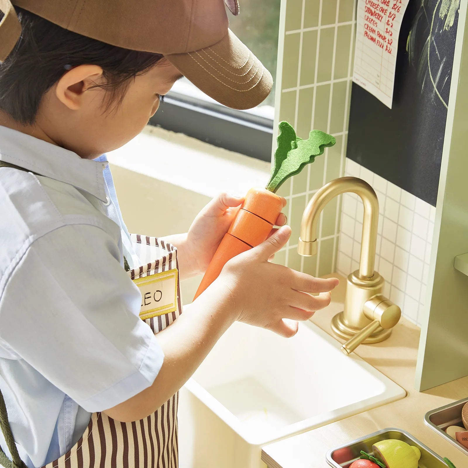 A child wearing a striped apron and cap enjoys pretend play with the Tiny Land® BistroSwitch Play Kitchen, featuring a golden faucet, kitchen accessories, and a basket of toy vegetables from Tiny Land.|Green