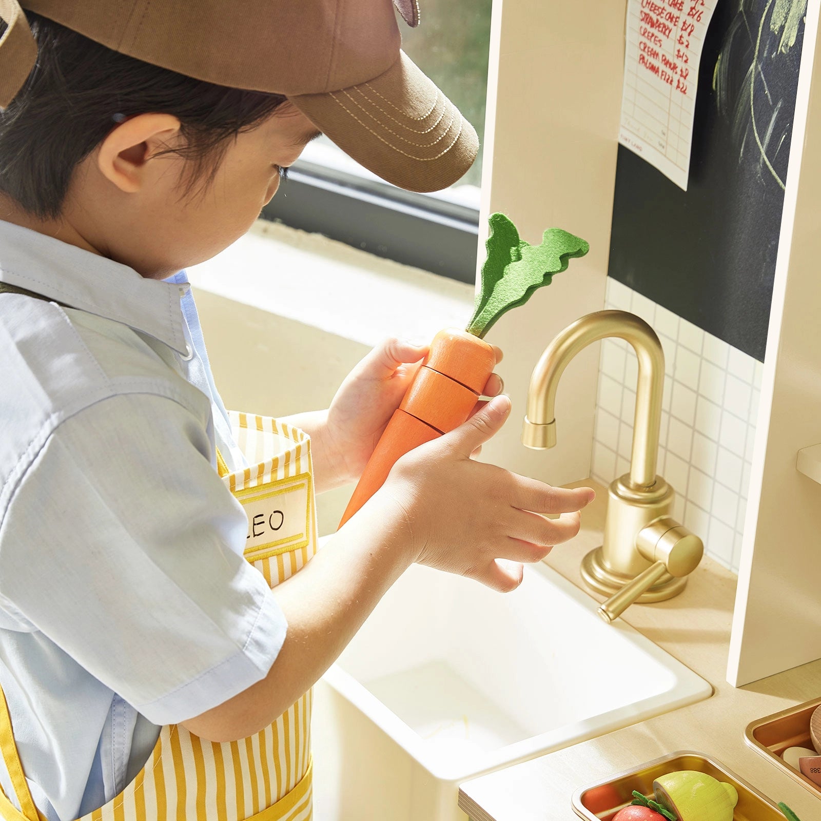 A child in a yellow apron stands by a window, enjoying pretend play at the Tiny Land® BistroSwitch Play Kitchen with toy fruits and vegetables from Tiny Land scattered nearby.|White