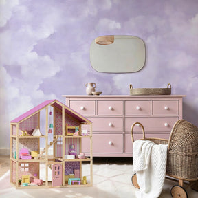 A pastel-themed child’s room featuring a pink dresser, Tiny Land® Sweetwood Unicorn Dollhouse & Wooden Dollhouse Dolls, a wicker bassinet with a white blanket, and a mirror on a cloud-painted purple and white wall.