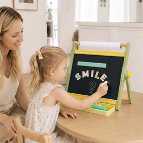 A woman and a young girl sit at a table, smiling. The girl writes SMILE on the Tiny Land® Double-Sided Magnetic Tabletop Easel by Tiny Land, drawing a cheerful flower in white on the chalkboard surface.|Green