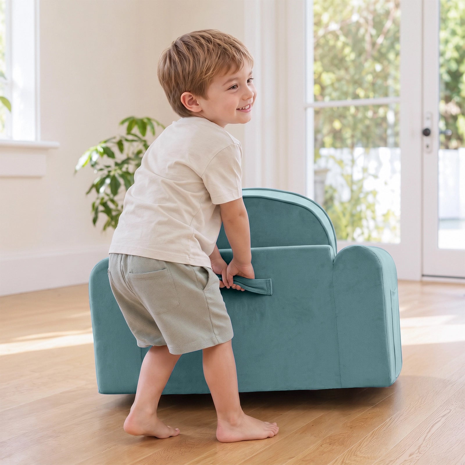 A young boy smiles while lifting a Tiny Land® GentleSnug Kids Sofa Chair—a small, light blue flip-open seat—in a bright room with wooden floors, large windows, and indoor plants.|Blue