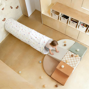 A young child crawls out of the Tiny Land® Adventure Play Tunnel - Colorful Polka Dots onto a padded play mat with soft blocks and scattered balls in a light, modern playroom.|white
