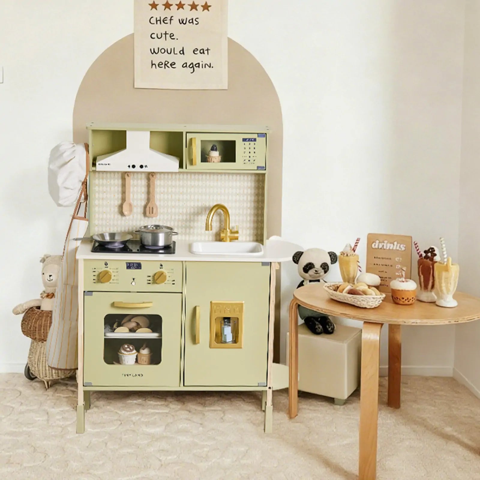 The Tiny Land® PopJoy Play Kitchen - Green by Tiny Land features a pastel green wooden kitchen with toy utensils, food, and a sink. Stuffed animals sit nearby, and a table holds play food. A wall sign reads, "Chef was cute. Would eat here again.