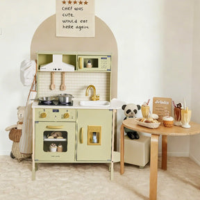 The Tiny Land® PopJoy Play Kitchen - Green by Tiny Land features a pastel green wooden kitchen with toy utensils, food, and a sink. Stuffed animals sit nearby, and a table holds play food. A wall sign reads, "Chef was cute. Would eat here again.
