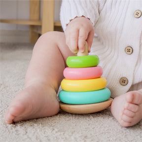 A baby in a white sweater sits on a carpet, stacking colorful rings from the Tiny Land® Montessori Early Skills Baby Set (7–12 months)—a classic Tiny Land toy that fosters fine-motor skills and sensory exploration.