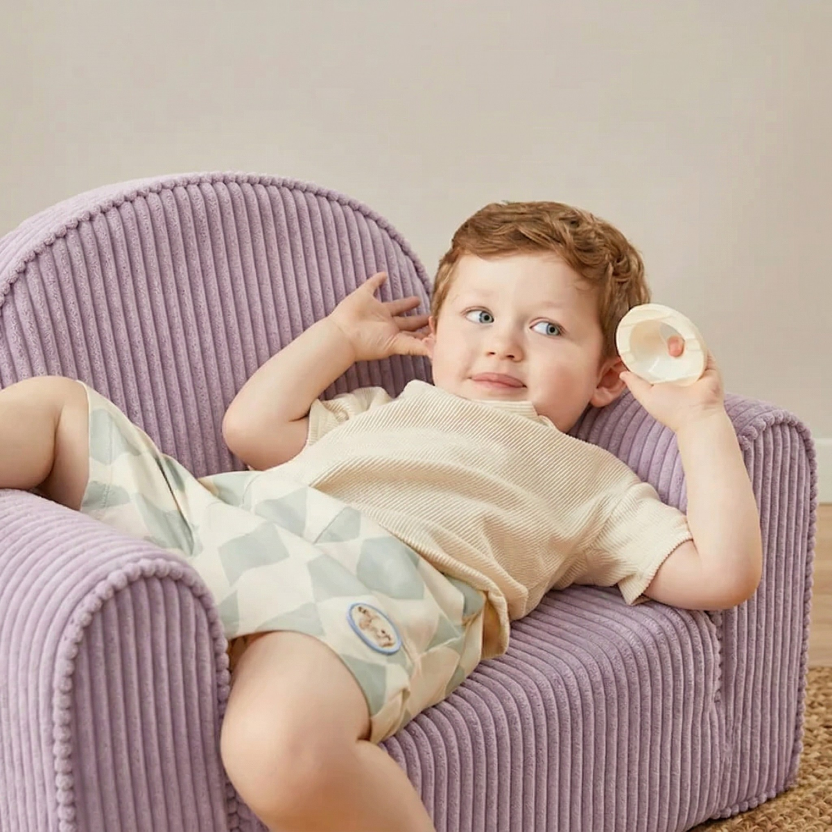 A young child with light brown hair lounges on the Tiny Land® GentleSnug Kid Chair in purple, holding a round translucent object. Relaxed and playful, the child wears a beige shirt and patterned shorts in this cozy setting by Tiny Land.