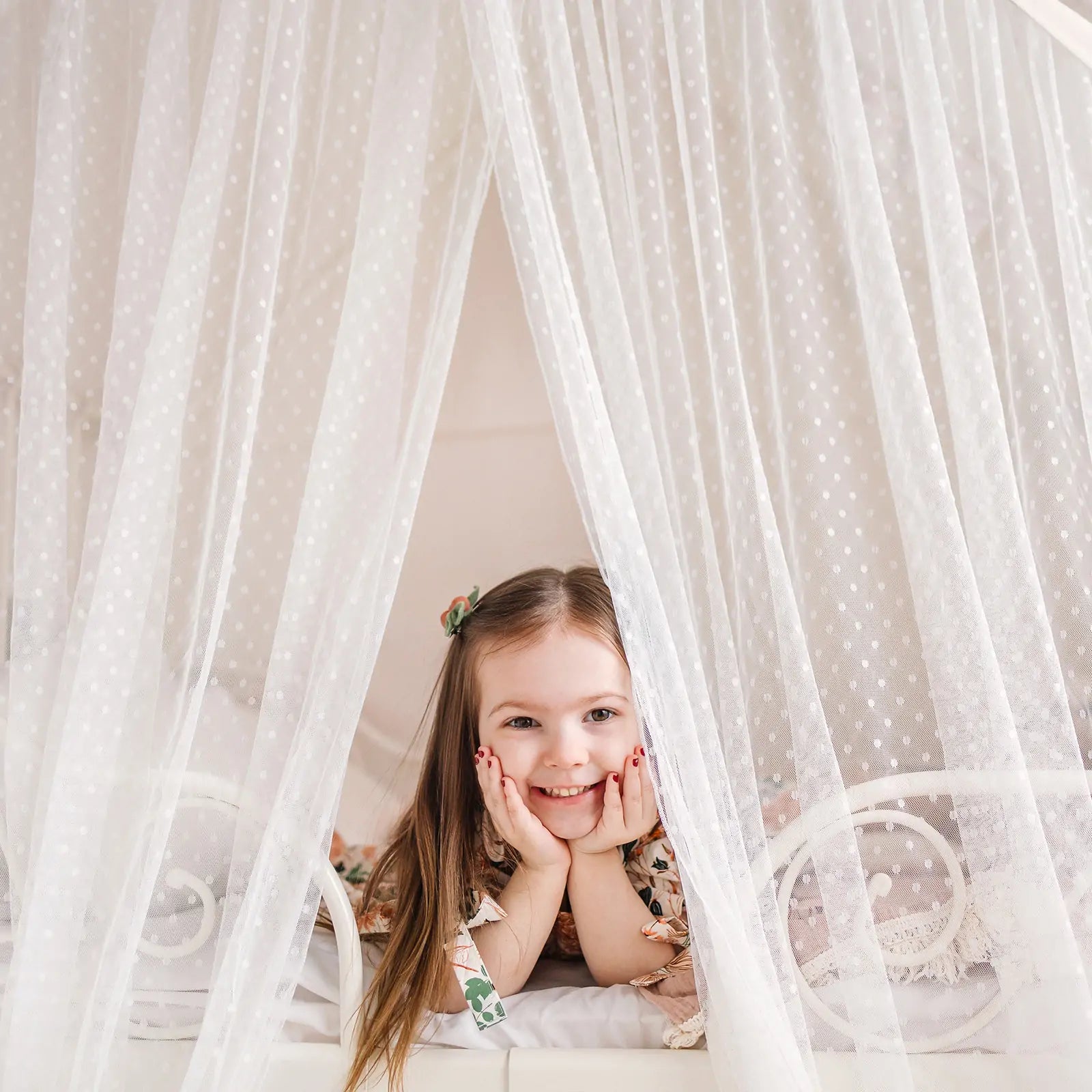 A young girl with long brown hair smiles as she rests on a bed inside the Tiny Land® Large Space Play House with Star Lights, surrounded by sheer white dotted curtains for a cozy, tent-like feel.