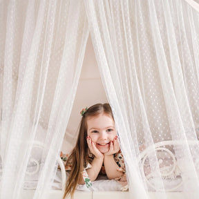 A young girl with long brown hair smiles as she rests on a bed inside the Tiny Land® Large Space Play House with Star Lights, surrounded by sheer white dotted curtains for a cozy, tent-like feel.